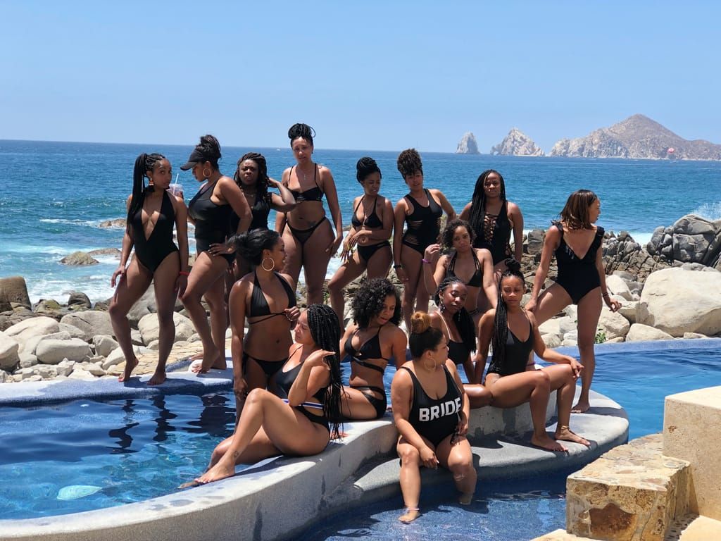 A bachelorette group in swimsuits by a villa pool with the Cabo San Lucas Arch behind them