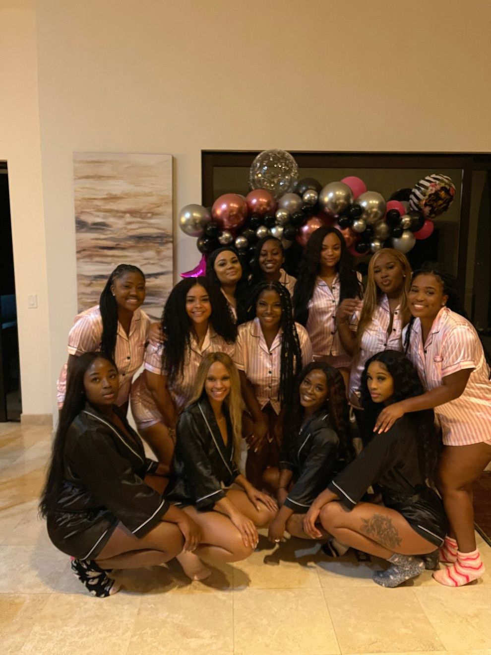 A bachelorette group in satin robes in front of a rose-gold balloon arch in their villa